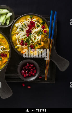 laksa with pumpkin and coconut milk, rice noodles, broccoli and pomegranate seeds in portioned black plates on a dark background. healthy vegan dish Stock Photo