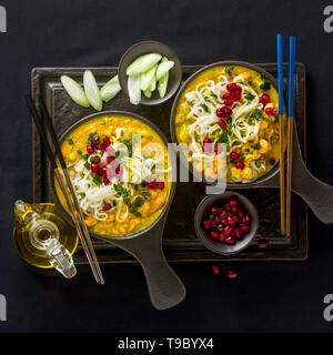 laksa with pumpkin and coconut milk, rice noodles, broccoli and pomegranate seeds in portioned black plates on a dark background. healthy vegan dish Stock Photo
