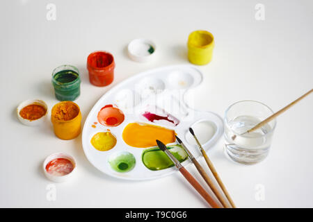Open jars with gouache of different colors: yellow, orange, red, green, as well as brushes, plastic palette and a glass of water for washing brushes. Stock Photo