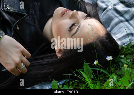 A positive woman lying down on a blanket in the meadow on spring sunny day and looking up and thinks. Girl with silk eyelashes Close up Stock Photo