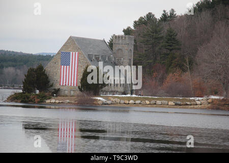 Old Stone church on edge of wachusett reservoir with large american flag hanging on the side with snow still on ground in May Stock Photo