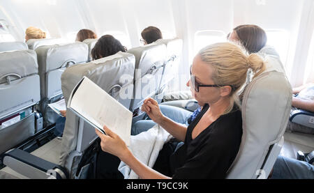 Woman reading magazine on airplane during flight. Female traveler reading seated in passanger cabin. Stock Photo