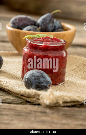 Jar of homemade plum jam with wooden bowl of juicy ripe fresh plums in background sitting on burlap sac. Stock Photo