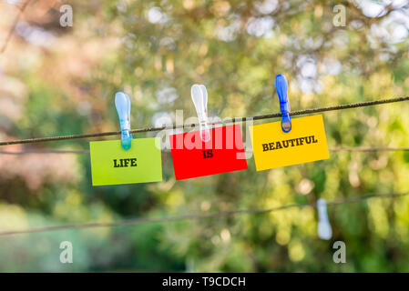 Life Is Beautiful concept with the words hanging on three green, red and yellow cards from a clothes line by plastic pegs over a leafy green nature ba Stock Photo