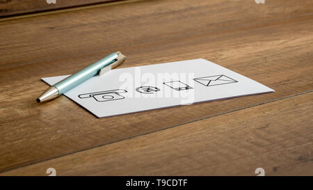 Ballpoint pen and four different contact icons printed on a white card or note lying on a wooden table. Stock Photo