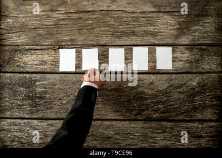 Top view of businessman hand placing five blank white peaces of paper or cards in a row on textured rustic wooden desk wit copy space ready for your t Stock Photo