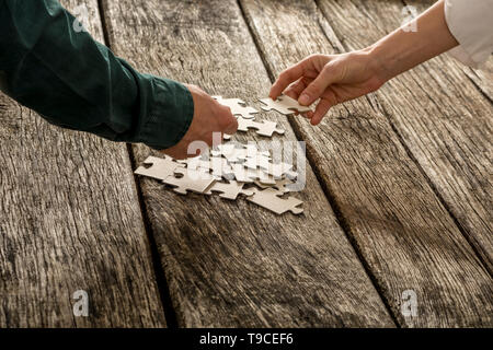 Two hands, male and female, matching two puzzle pieces over a pile of other pieces lying on a textured rustic wooden desk. Conceptual of teamwork and  Stock Photo