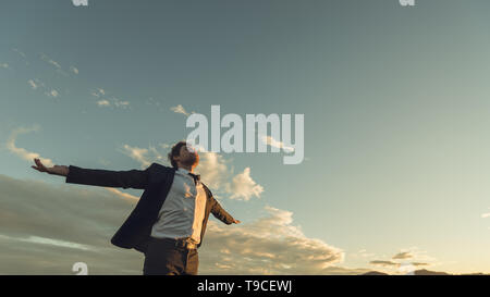 View from below of a young businessman in a suit enjoying his freedom and professional independence with arms wide open standing under evening sky. Stock Photo