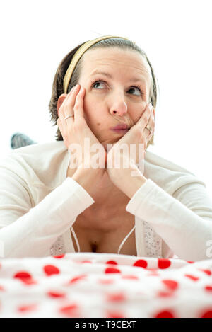 Young woman lying thinking on top of a colorful red polka dot counterpane on a bed looking up into the air with a contemplative expression. Stock Photo