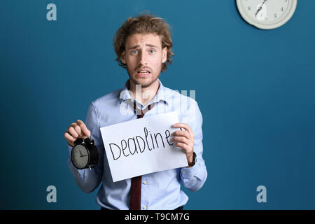 Stressed businessman holding sheet of paper with word DEADLINE and alarm clock on color background Stock Photo
