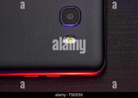 A fragment of the cell phone case in black with red backlight. The phone lies on the black technological surface of the camera up with red lights. Stock Photo