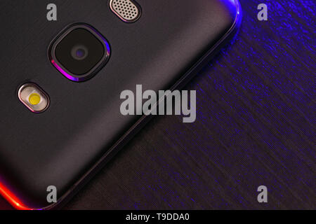 A fragment of the cell phone case in black with blue and red backlight. The edge of the phone lies on the black technological surface of the camera up Stock Photo