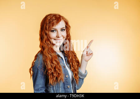 Redhead beautiful yong woman smiling showing product. Studio portrait of attractive girl with curly hair pointing to the side on orange background wit Stock Photo