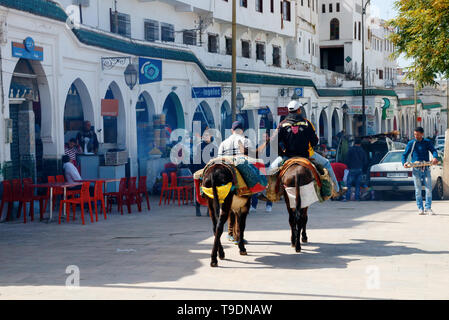 Main square of Moulay Idriss with shops, bars and two men with donkeys walking along the arcade on a sunny day. Moulay Idriss Zerhoun, Morocco Stock Photo