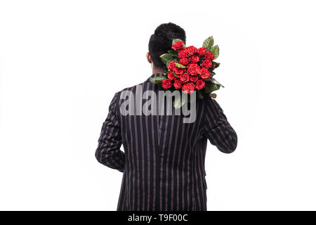 Handsome yound man in suit is standing with red roses behind the back on grey background. Stock Photo