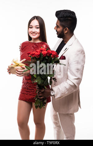 Beautiful romantic asian couple, young asian woman in dress holding red roses and handsome indian man in suit are in love isolated on white background Stock Photo