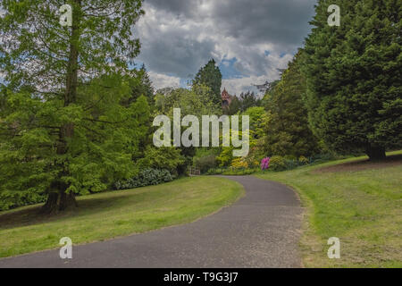 Pathway in between the grass hills at Belle Vue park, Newport, Wales UK with the pavilion nestled in the trees. Dry sunny day. Dark cloudy sky. Stock Photo