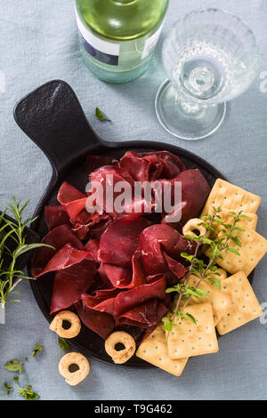 Italian bresaola served sliced on a tray on a table with white wine, crackers, grissini and taralli with aromatic herbs on a blue linen festive tablec Stock Photo