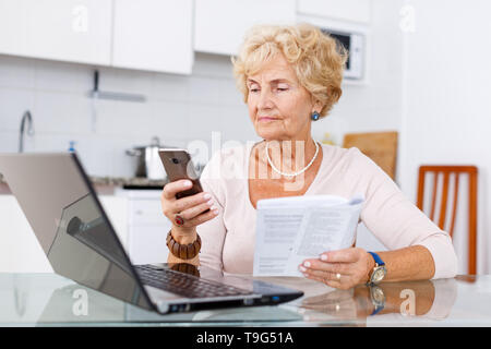 Active senior woman making order by phone using laptop in her kitchen Stock Photo