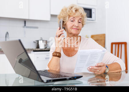 Active senior woman making order by phone using laptop in her kitchen Stock Photo