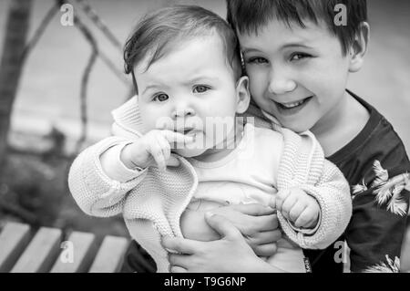 Elementary school age Caucasian child boy holding tenderly his beloved little sister girl somewhere outside. Brother to sister sincere love concept Stock Photo