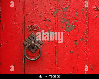flaking red paint on old solid timber barn door with shiny cast iron ring door handle on ornate diamond pattern fixing plate in Cumbria,England UK Stock Photo