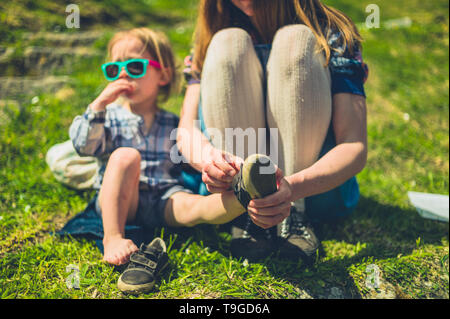 A young mother is helping her toddler put on his shoes in the park on a sunny summer day Stock Photo