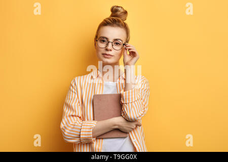 clever cute awesome woman holding a book, touching her glasses and posing to the camera. isolated yellow background, studio shot. Stock Photo