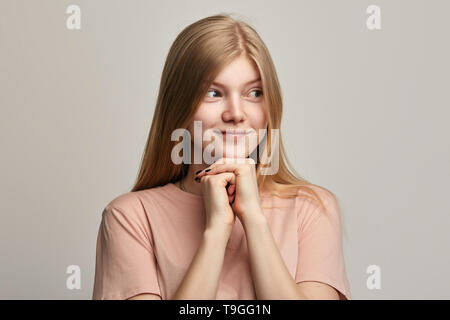 Sly beautiful girl planning evil trick, holding hands in front of her chest and smiling mysteriously. Pensive cunning young woman with fists in front of her looking aside. Stock Photo