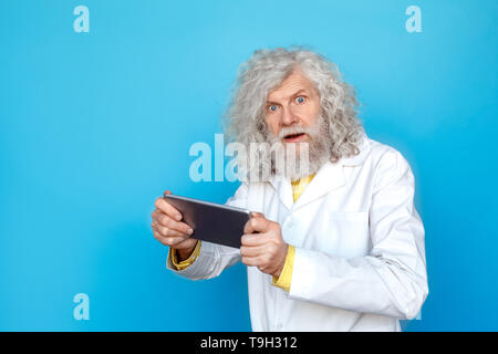 Old long-haired man wearing doctor's gown studio isolated on blue wall standing holding digital tablet watching online video looking camera surprised Stock Photo