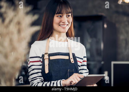 Asian female barista wear apron hold tablet computer coffee menu with smiling face in cafe waiting order from customer ,Coffee business owner concept. Stock Photo