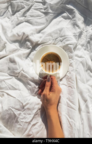 Womans hand holding a cup of coffee on the bed with white linen on the room with natural sunlight, top view Stock Photo