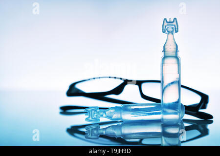 Artificial tears eye drops encapsulated in plastic pipettes with eye glasses reflected on glass table with blue background. Horizontal composition. Fr Stock Photo