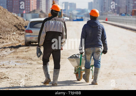 Two workers in helmets drive a wheelbarrow against the background of new buildings. Stock Photo
