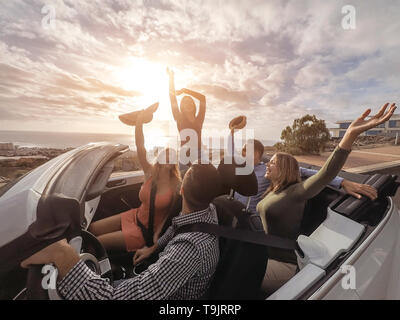 Happy friends with their hands up having fun in cabriolet car on vacation - Young people laughing and enjoying together during travel road trip Stock Photo