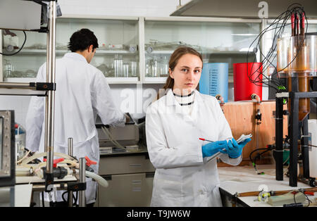 Portrait of young glad  friendly woman lab technician writing report on results of chemical experiments in notebook Stock Photo
