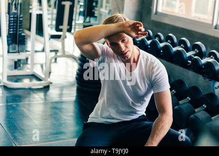 Young man wiping sweat off forehead with dumbbells and fitness equipment inside gym. He is tired and exhausted from weightlifting and weight training. Stock Photo