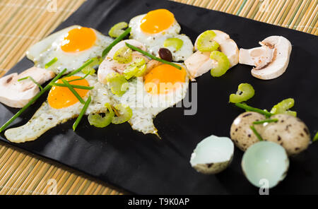 Tasty fried quail eggs with raw champignons and fresh greens on black plate Stock Photo