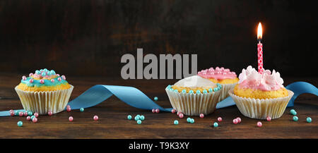 pink and blue decorated muffins or cupcakes with a ribbon and a burning candle on a rustic wooden board against a dark brown background, panoramic for Stock Photo