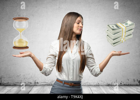 Young business woman holding sand glass and bundles of dollars in her hands on grey wall background Stock Photo