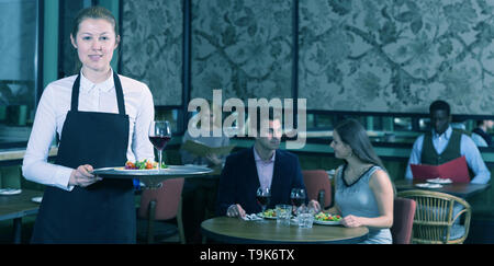 Polite waitress wearing apron holding tray with dishes in restaurant ...
