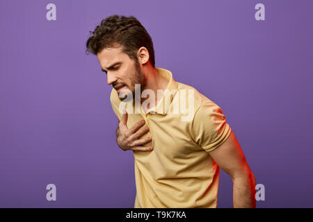 weak sick guy having problems with heart. attractive unhappy man in yellow T-shirt touching his chest, heart. close up photo. isolated blue background.myocardial infaction. heart palpitations Stock Photo