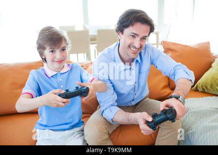Father and son sitting on sofa in lounge playing video game Stock Photo