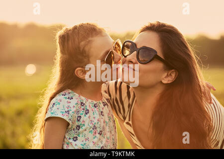 Happy fashion kid girl whispering her mother the secret in trendy sunglasses in profile view and looking on nature background. Closeup portrait of hap Stock Photo