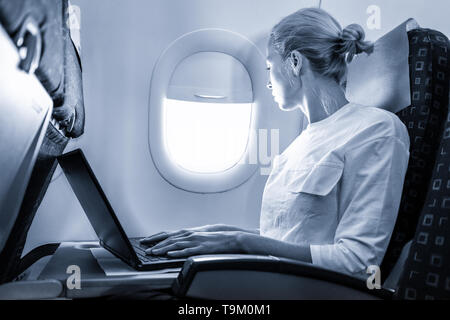 Attractive caucasian female passenger looking through the plain window while working on modern laptop computer using wireless connection on board of Stock Photo