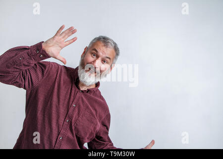 Adult gray-haired man with a beard in a red shirt smiles and waves his arms against a white wall. Stock Photo