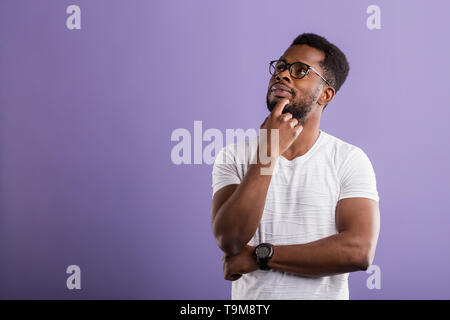 Dark skinned unshaven man with short curly hair in casual outfit holding chin with hand, looking up with dreaming expression, smiling, thinking about  Stock Photo