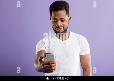 Studio shot of cheerful smiling african american unshaven young man holding cellphone, taking selfie portrait, making video call, wearing white clothe Stock Photo