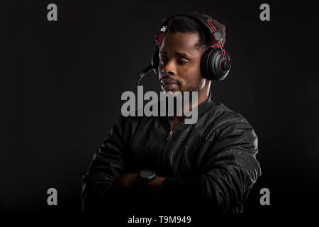 Portrait of dark skinned male with beard listening attentively audiobook or song on headphones, wearing black jacket looking pensively down, standing  Stock Photo