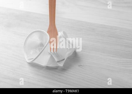 White plastic cup crumpled with a wooden eco fork on the wood table. Part of the image is black and white and part is colored. The concept of plastic  Stock Photo